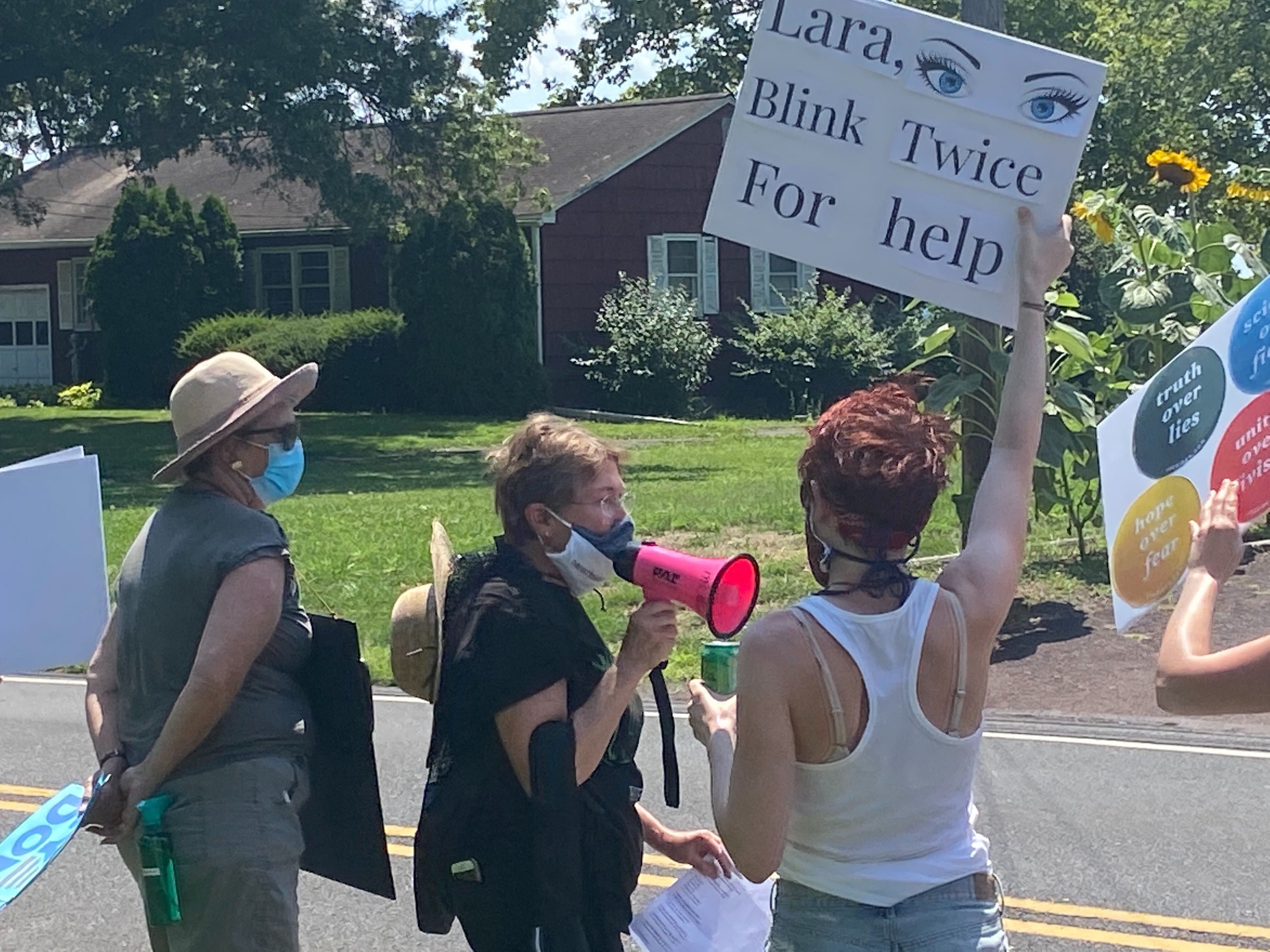 Three white protesters wearing face masks: one holds up a sign reading &quot;Lara, blink twice for help,&quot; another is speaking into a bullhorn. 