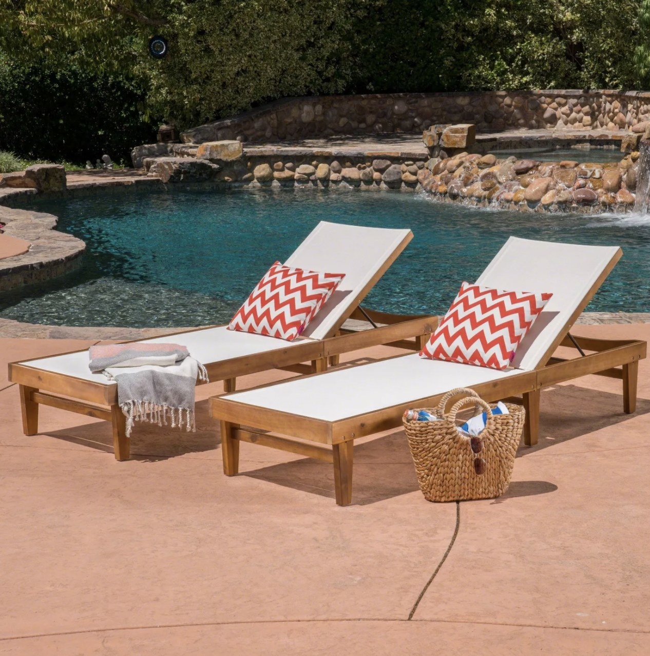 A pair of chaise lounges made of acacia wood and white mesh with chevron accent pillows in front of a pool
