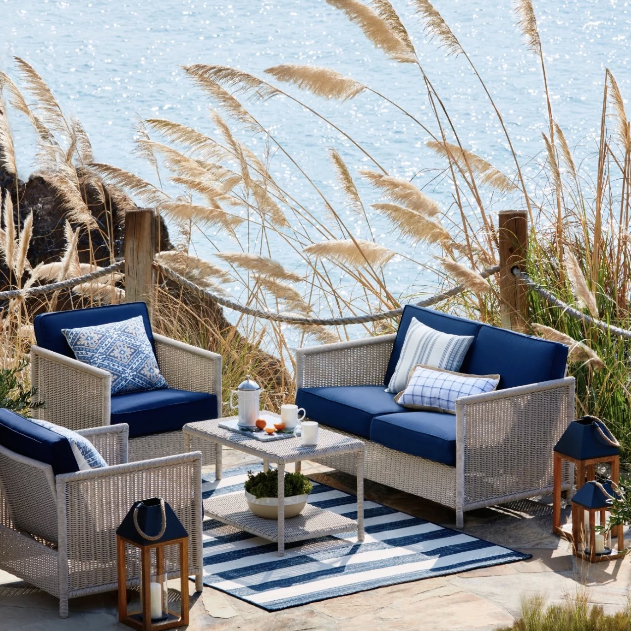 A lightly colored wicker loveseat with blue cushions and two matching armchairs and a coffee table on a patio by the water.
