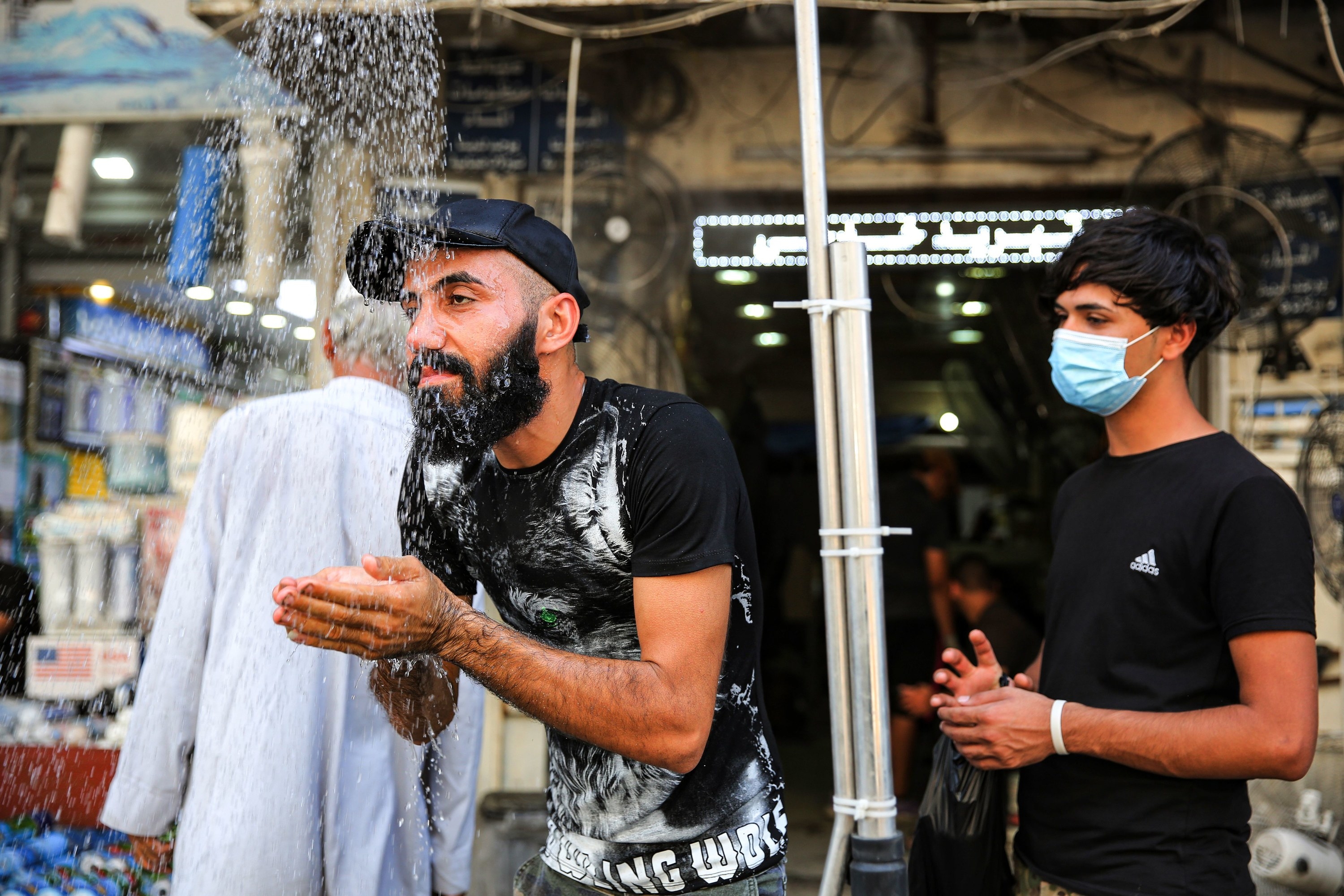A man cups his hands under a spray of water while a young man wearing a face mask stands behind him
