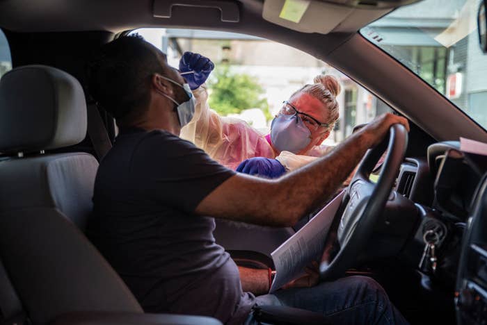 A medical professional, wearing a face mask, reaches an arm into a car and inserts a swab into the driver's nose