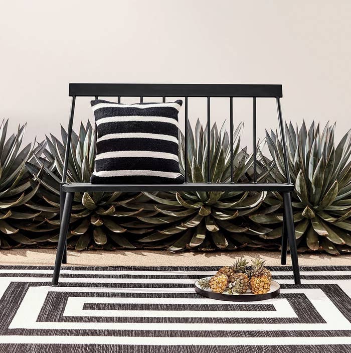 A large rug in a black and white mitre-striped pattern underneath a black bench and in front of a garden of large succulents