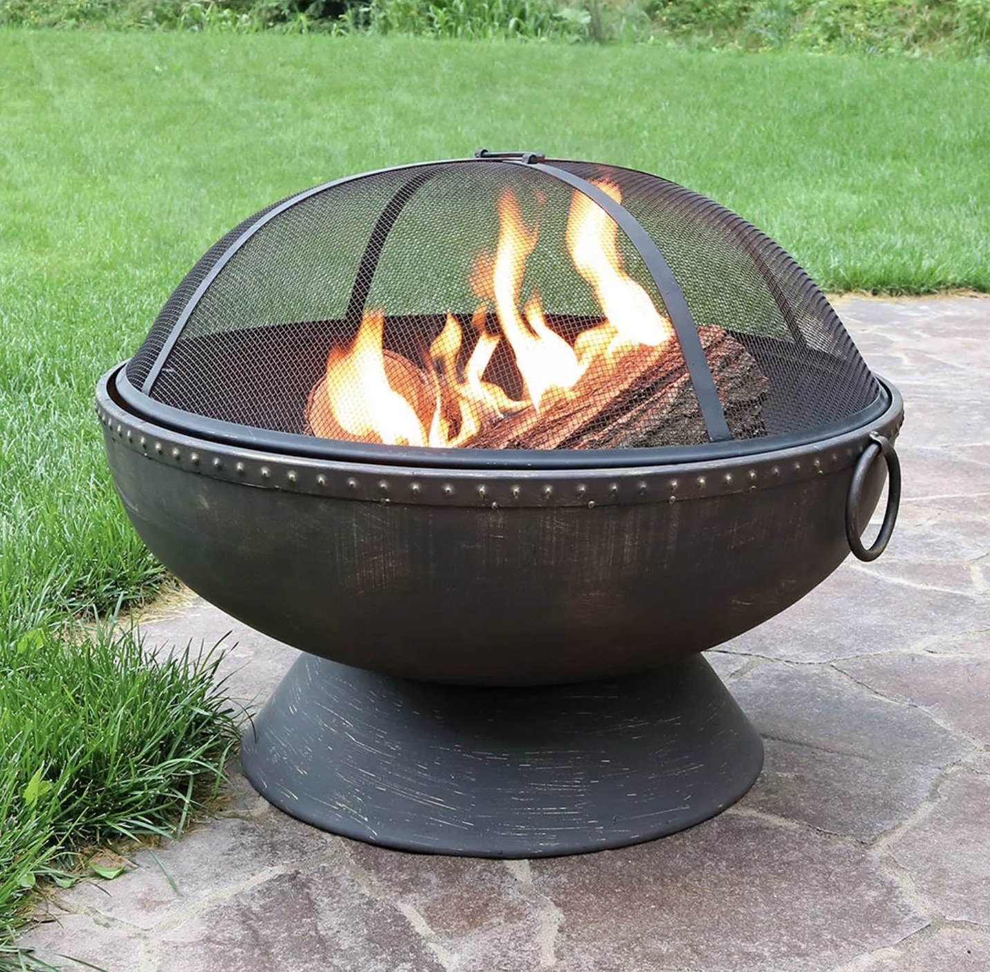 A fire pit bowl with a brushed steel pedestal base with a brass-tack edge, rings, and a domed screen covering a fire