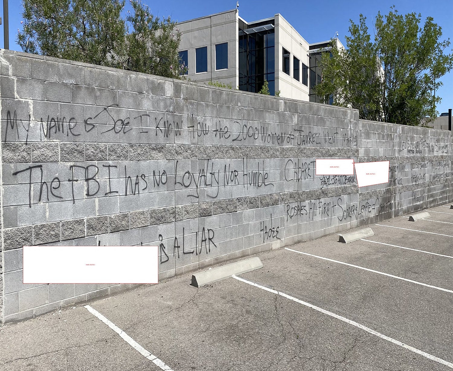 Black graffiti on a wall next to a parking lot reads "The FBI has no loyalty nor humble" among other phrases, while some words are redacted