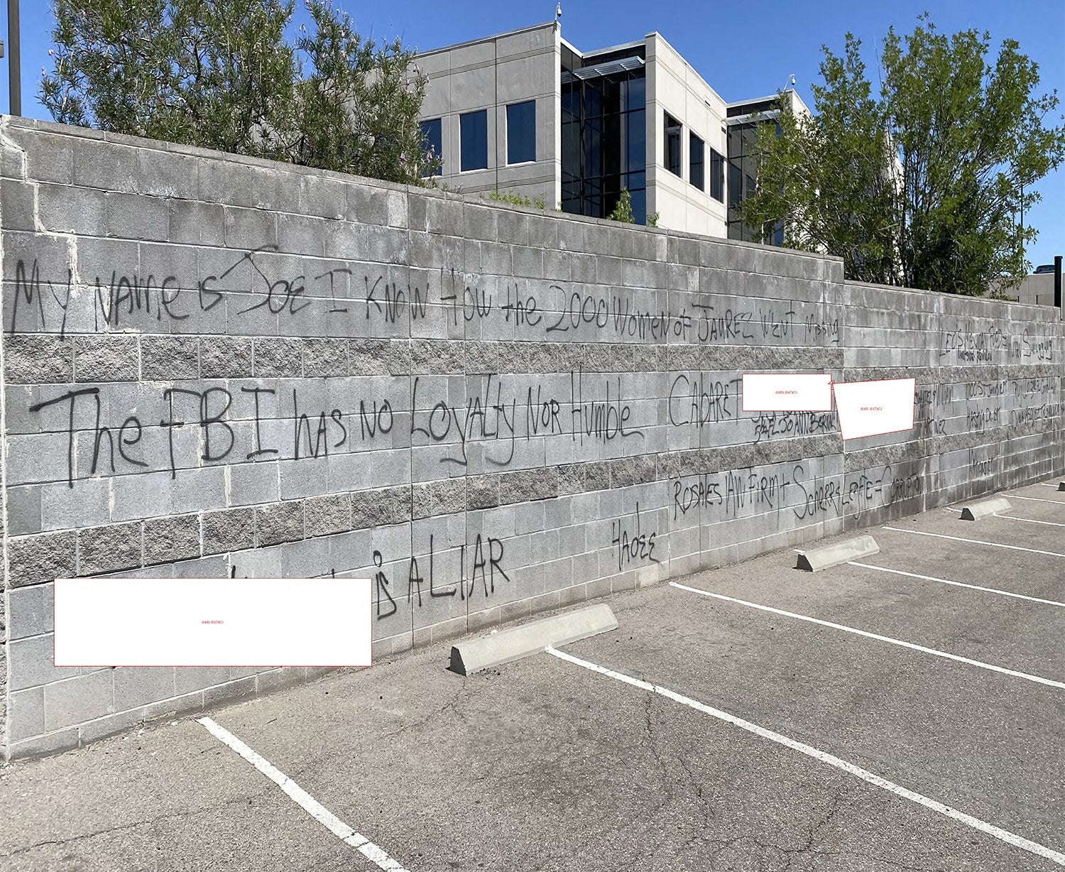 Black graffiti on a wall next to a parking lot reads &quot;The FBI has no loyalty nor humble&quot; among other phrases, while some words are redacted