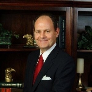 Al Hartman wears a suit, tie, and pocket square as he smiles in front of a book case