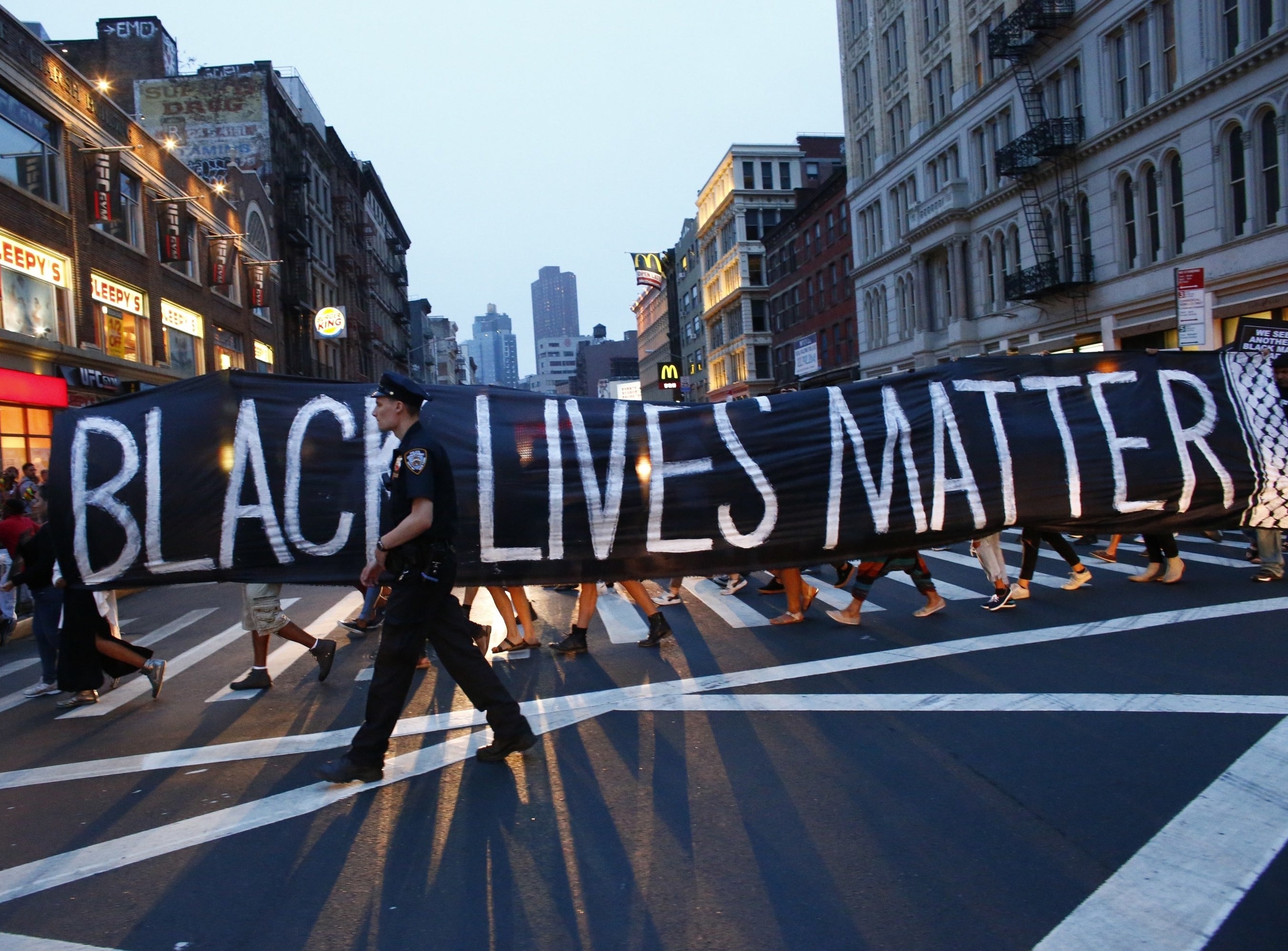 A large banner that says, &quot;Black Lives Matter,&quot; held by protestors across the length of a street as a police officer walks in front of it