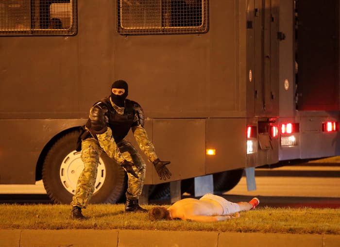 A man in military-style gear and a mask gestures to a man lying shirtless, facedown on the ground.