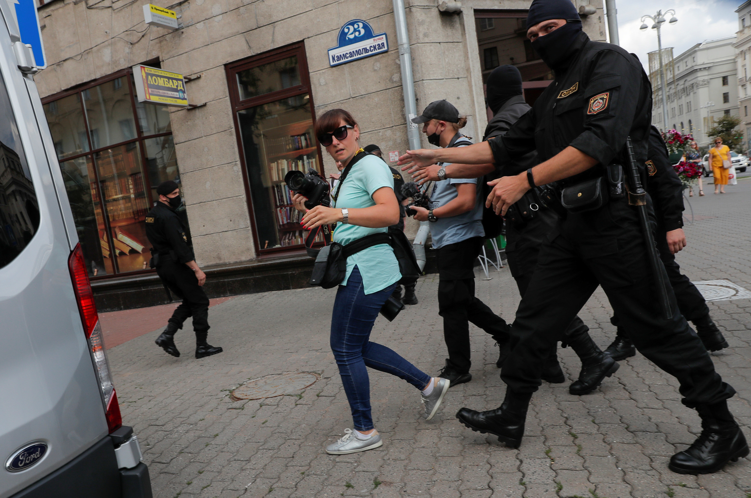 Officers wearing face masks approach a woman with a camera and other professional gear.