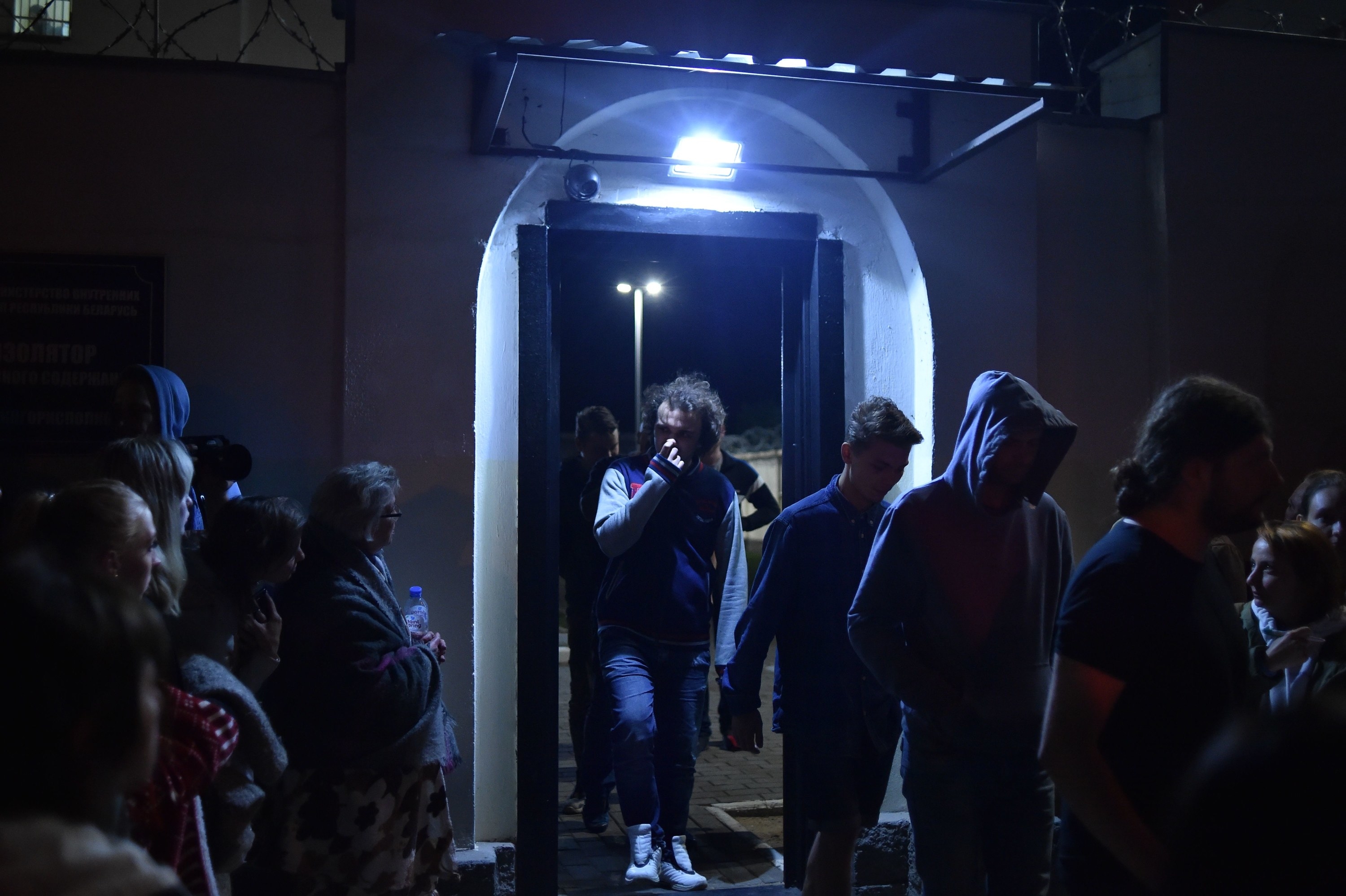 A line of people, many with their heads bowed, exit a prison doorway to crowds of people waiting outside.