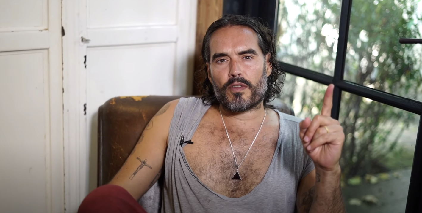 Russell Brand points his finger up while sitting in a chair at his home