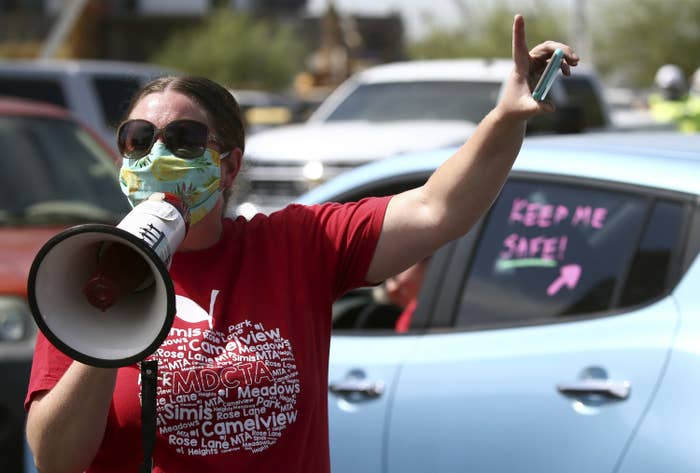 A teacher wearing a mask uses a megaphone while protesting amid cars, one of which has a window sticker that says keep me safe