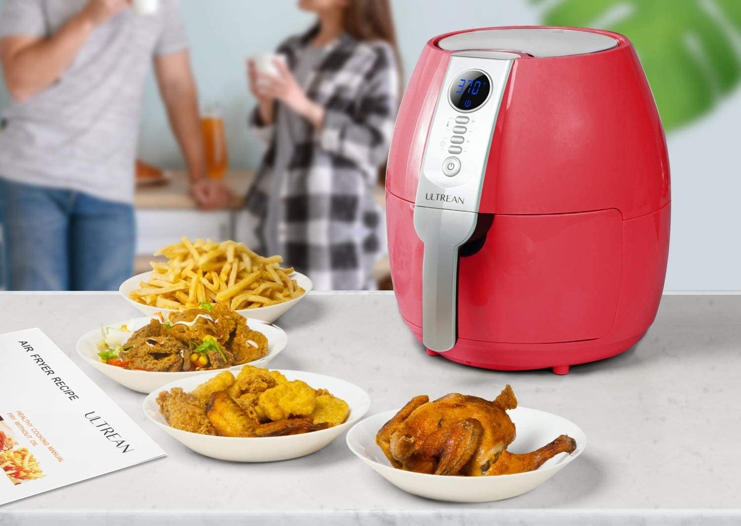 The red air fryer on a counter next to plates of fries, chicken, and other foods