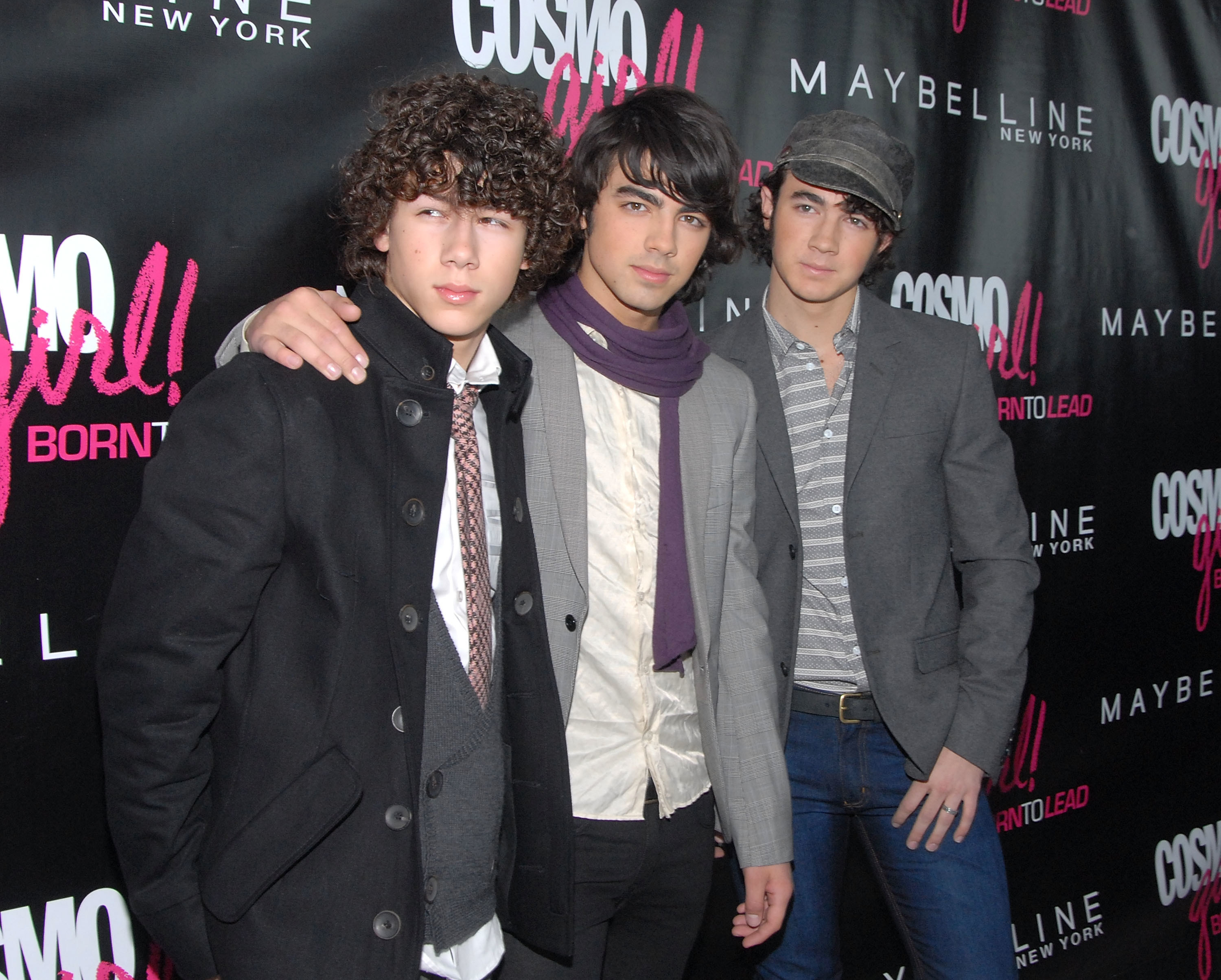 Nick (in black coat and tie), Joe (in a ivory silk shirt, grey jacket, and purple scarf), and Kevin Jonas (in a grey jacket, grey stripped shirt, and old fashion hat) on the red carpet of a CosmoGirl.