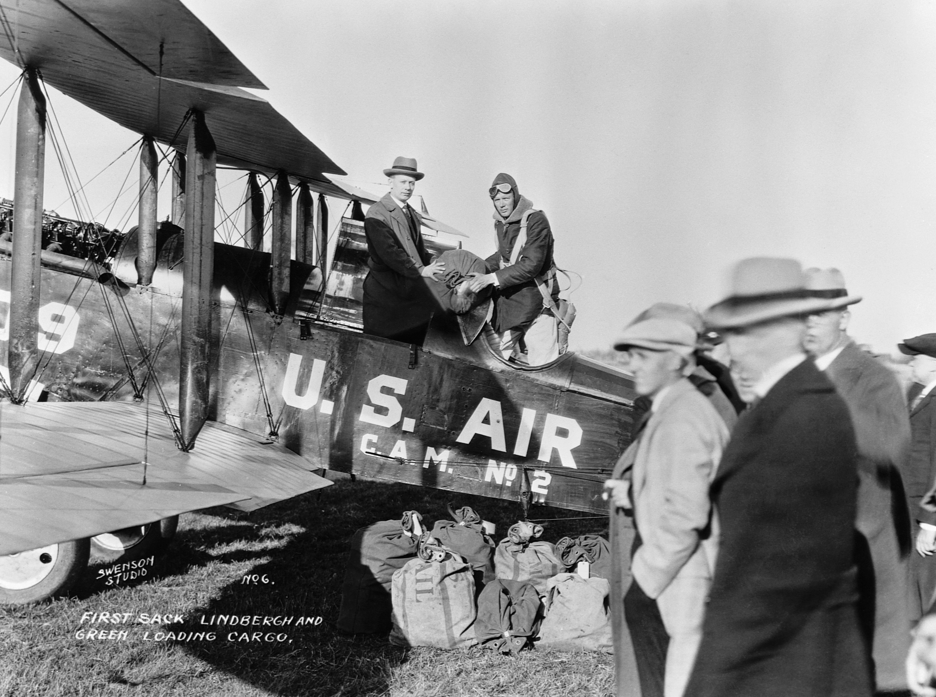 Two men, one in a fedora and one in a soft aviator helmet, stand holding a bag of mail in the cockpit of an old plane that says US Air