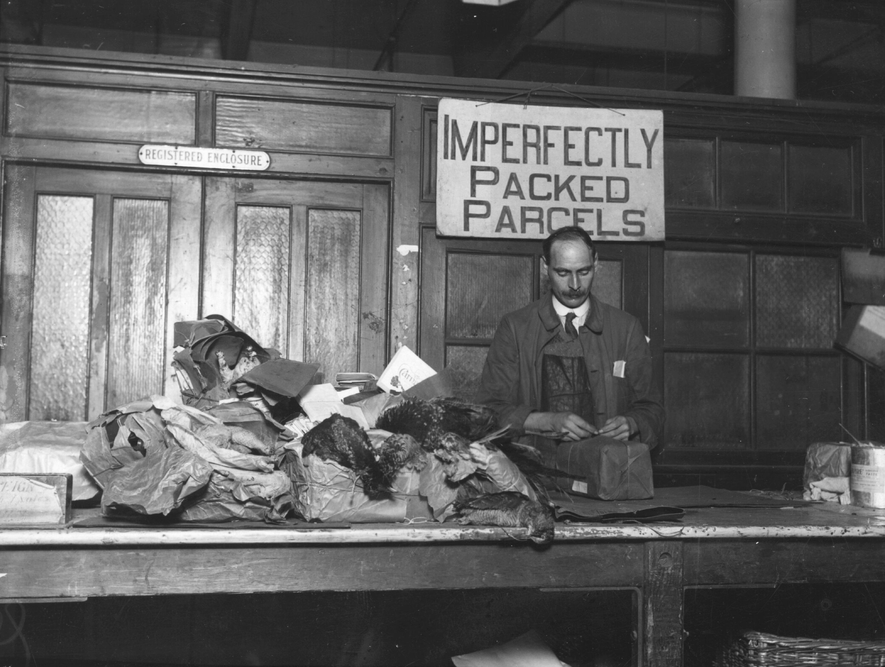 A man in front of a pile of messy packages stands under a sign reading &quot;imperfectly packed parcels&quot;