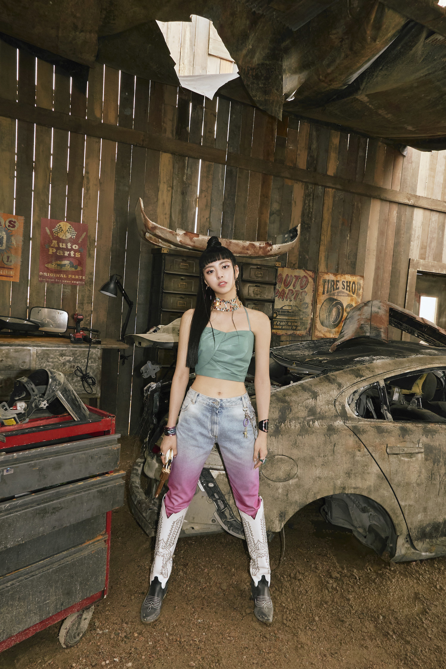 Yuna stands in front of a destroyed car in a country western-themed workshop 