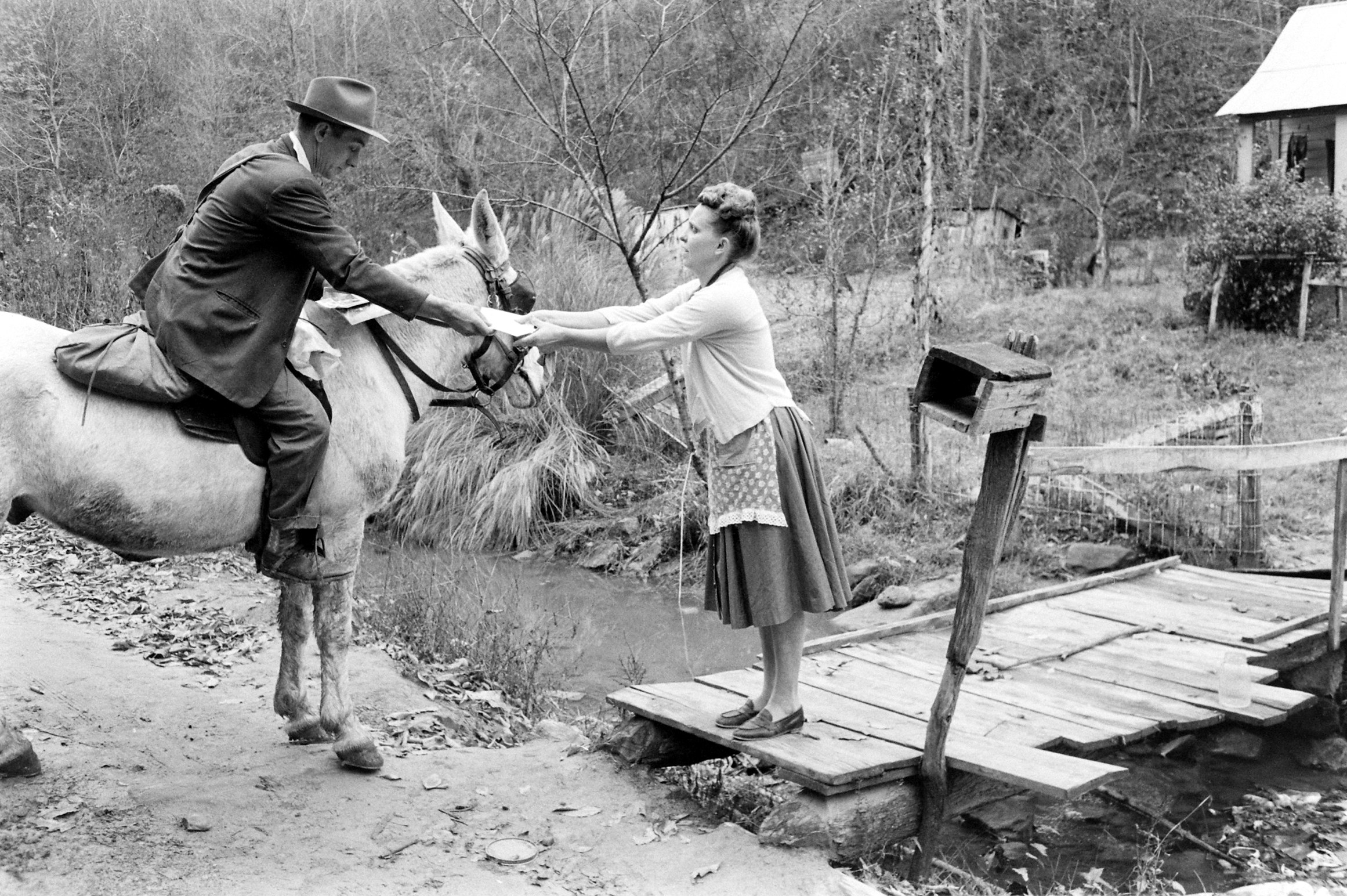 A man on horseback hands a letter to a woman in a dress and apron standing on a wooden bridge