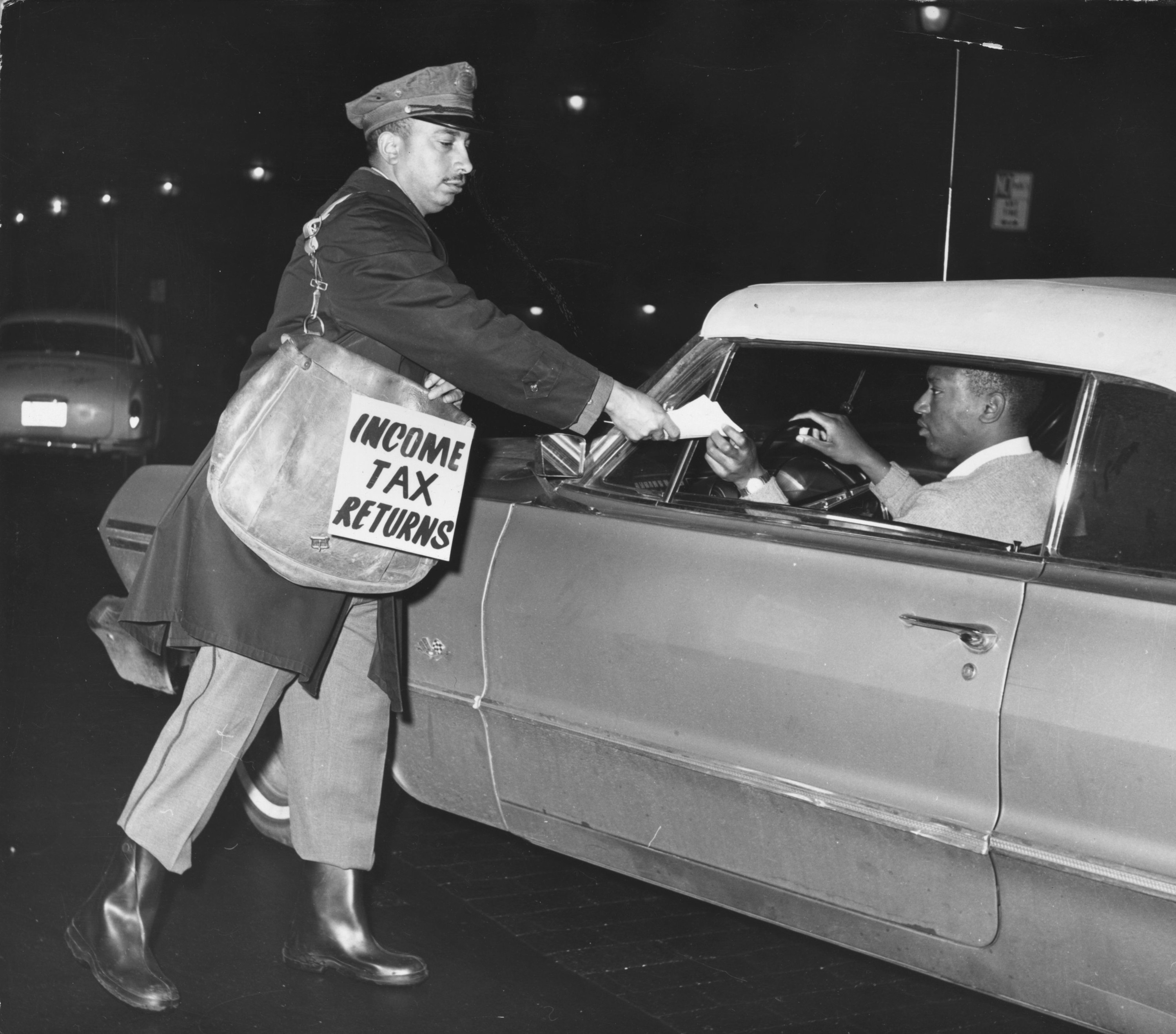 A white man with a shoulder bag reading &quot;income tax returns&quot; takes a piece of paper from a black man driving a car