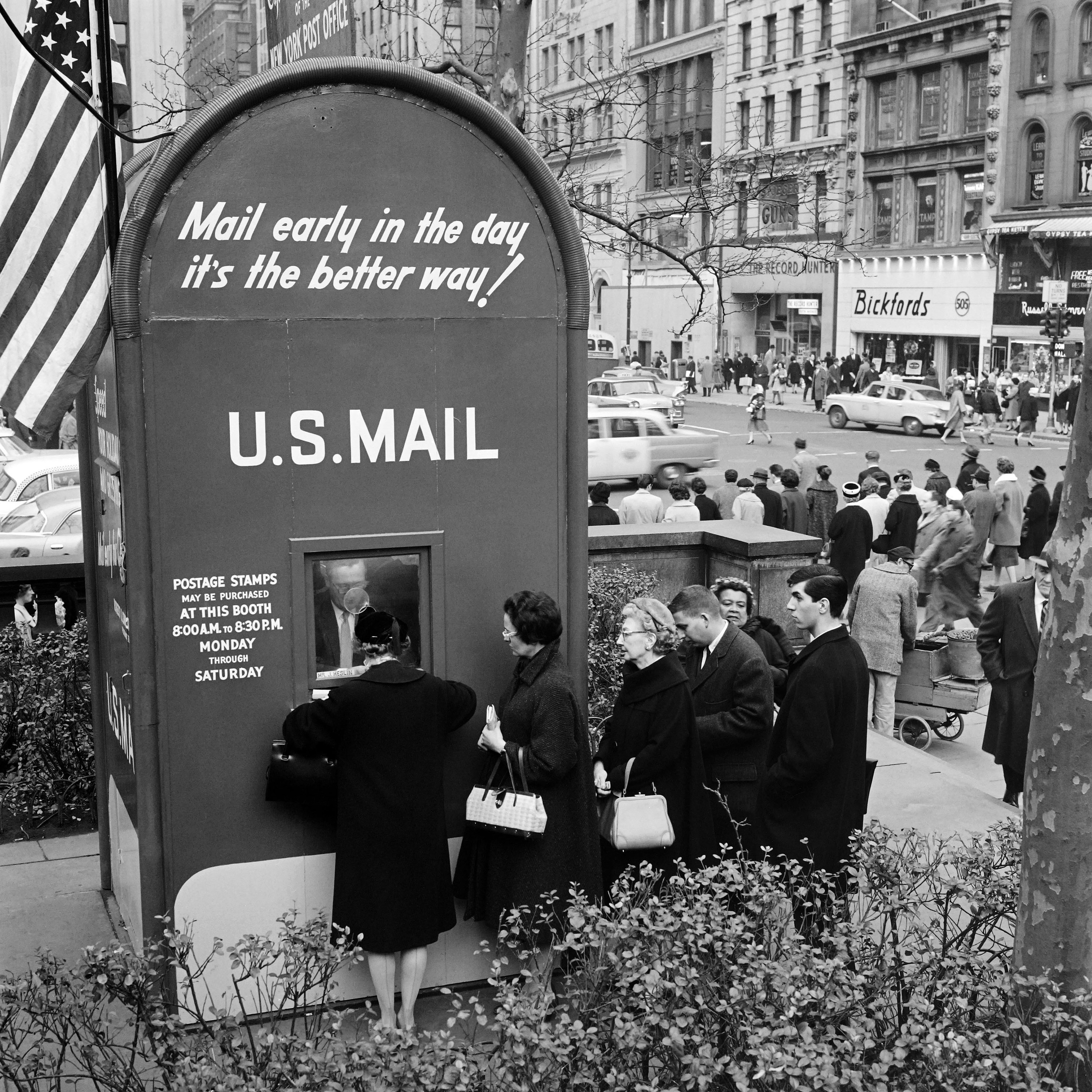 Well-dressed men and women stand in line to drop off letters in a booth shaped like a giant mailbox in a city square