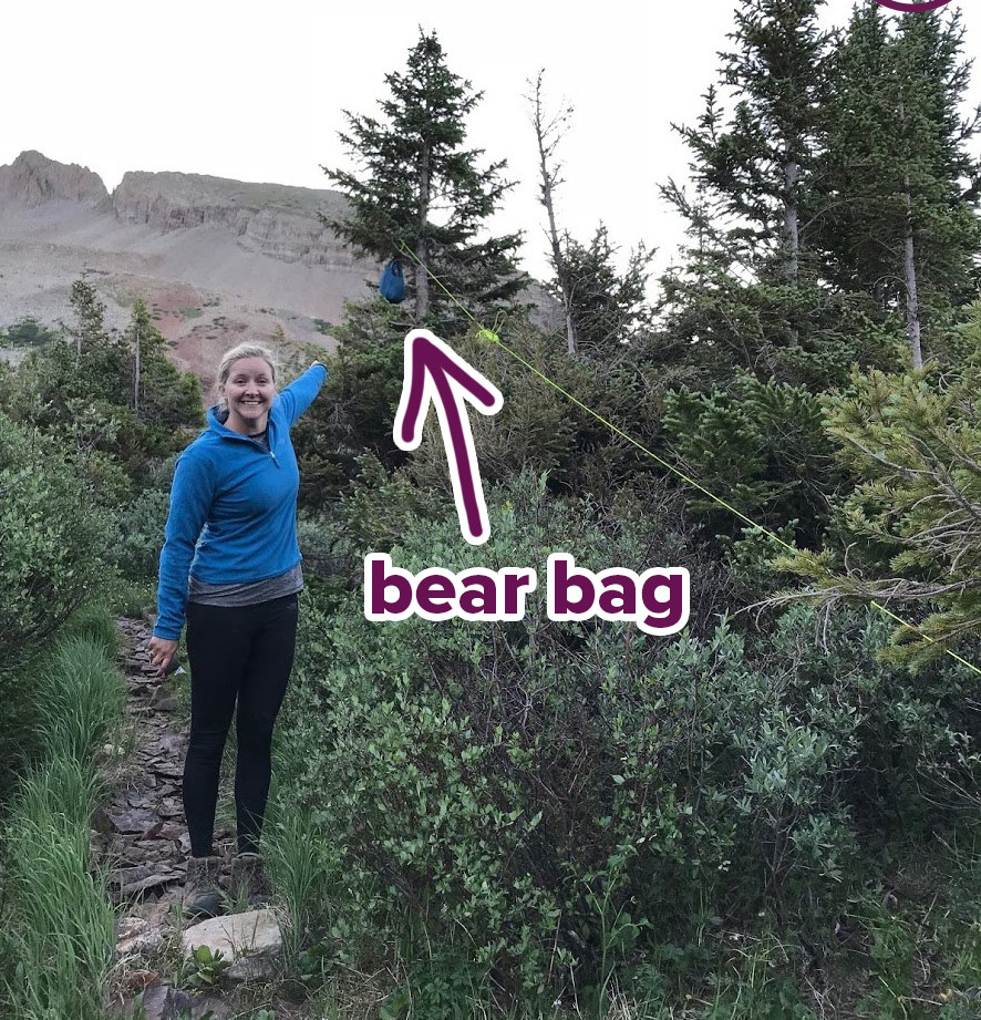 woman on a rugged path pointing towards a small bear bag hanging in the trees