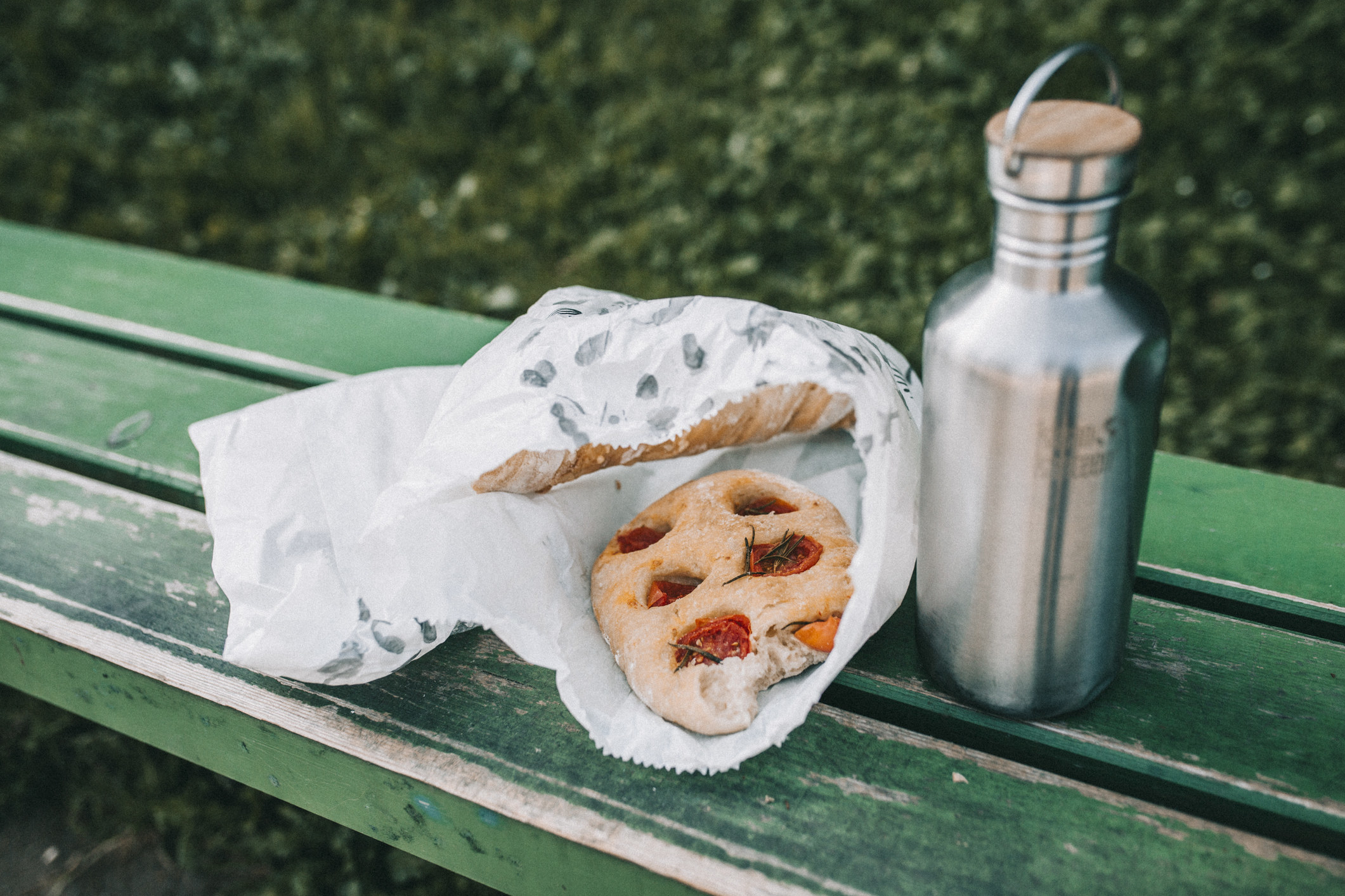 tomato focaccia in a paper bag next to a metal drink bottle, sitting on a wooden bench outdoors