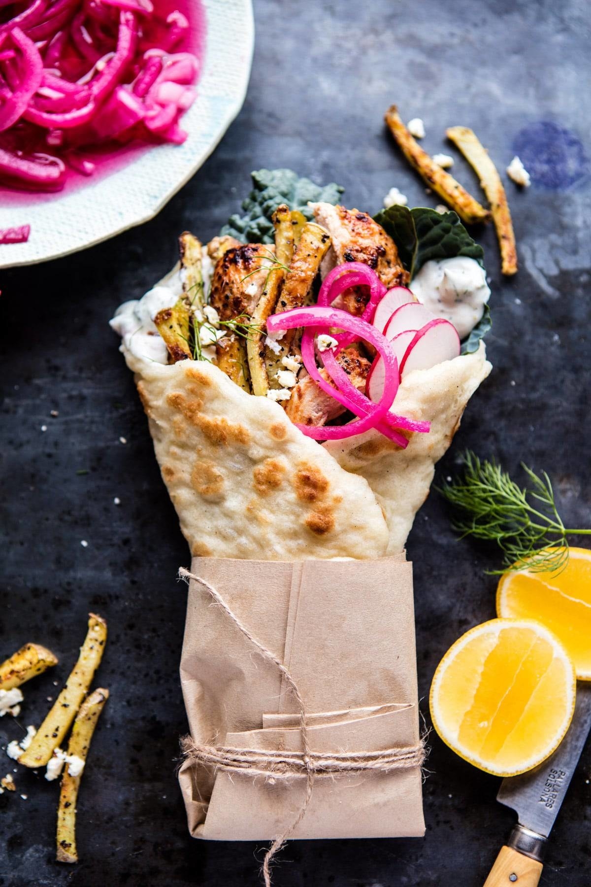 A fluffly pita wrapped in paper holds roasted chicken, bright pickled onions, crunchy radish, fries, and crumbled feta