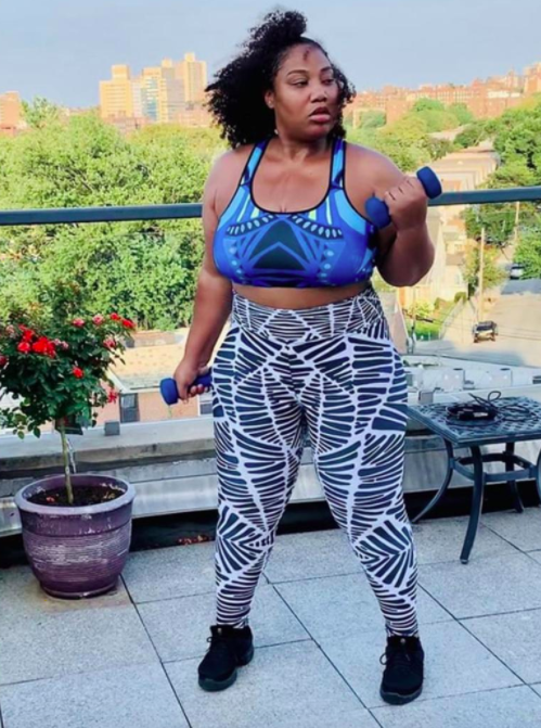 Model wears black-and-white-patterned high-rise leggings, a colorful sports bra, and black sneakers while lifting blue weights on a balcony
