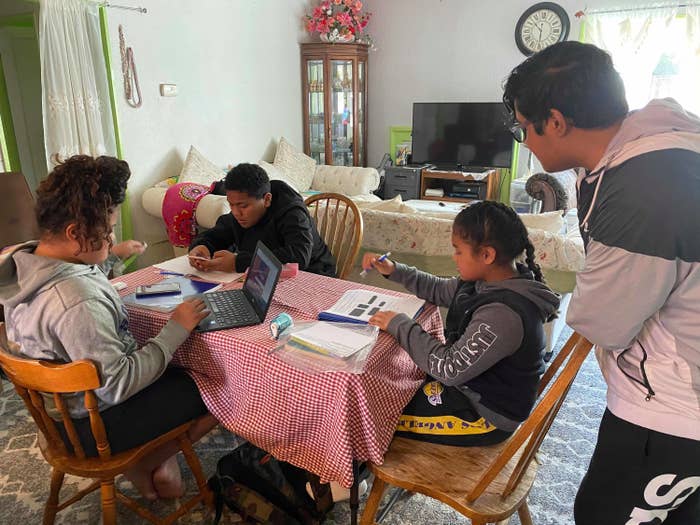 Three young people sit around a table working on schoolwork, while one person stands over them