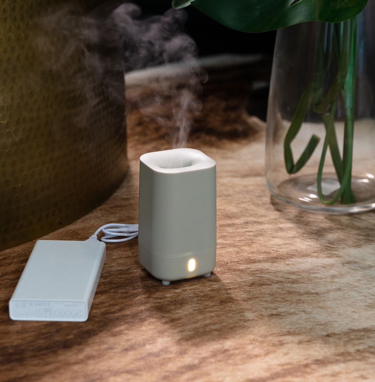The small diffuser sitting on a table with mist coming out of the top and a battery pack sitting next to it