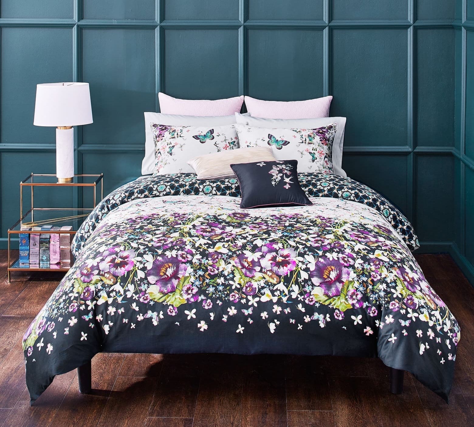 Floral comforter in navy with purple and green flowers all over it on a bed with pillows with floral and butterflies on them