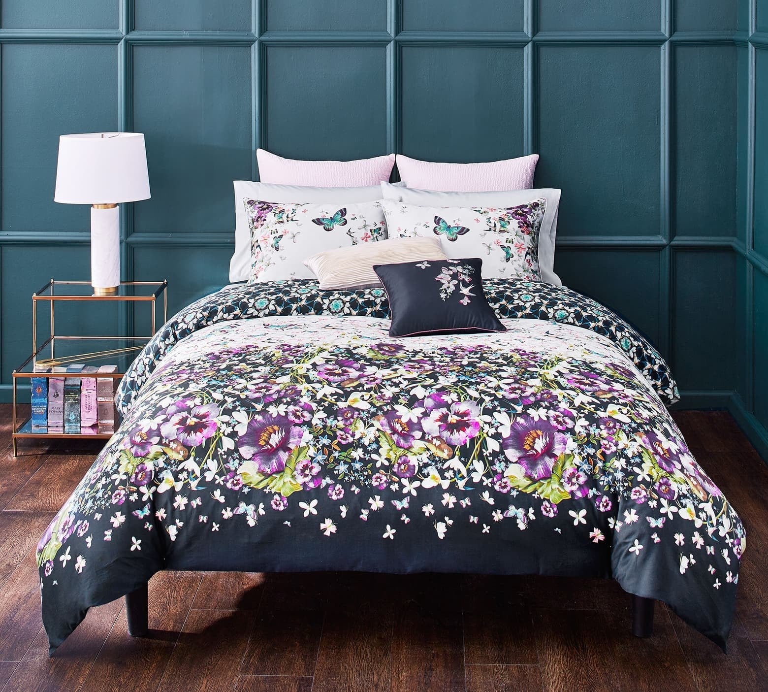Floral comforter in navy with purple and green flowers all over it on a bed with pillows with floral and butterflies on them