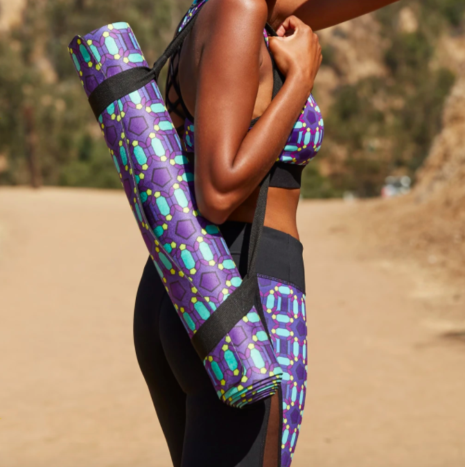 Model carries rolled-up blue, yellow, and purple-patterned yoga mat in a black strap across their shoulder