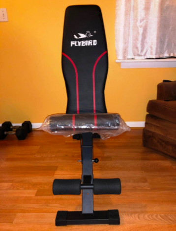 Reviewer shows black and red workout bench in front of a stack of black dumbbells in their living room
