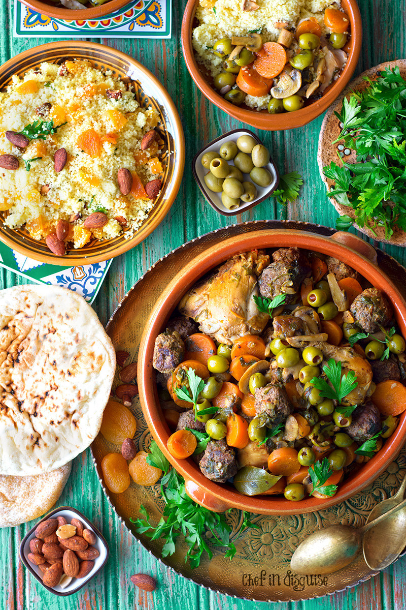 An orange tagine filled with roasted chicken, meatballs, olives, and carrots on a decorative plate and surrounded by side dishes, like fluffy cous cous, browned pita, and almonds