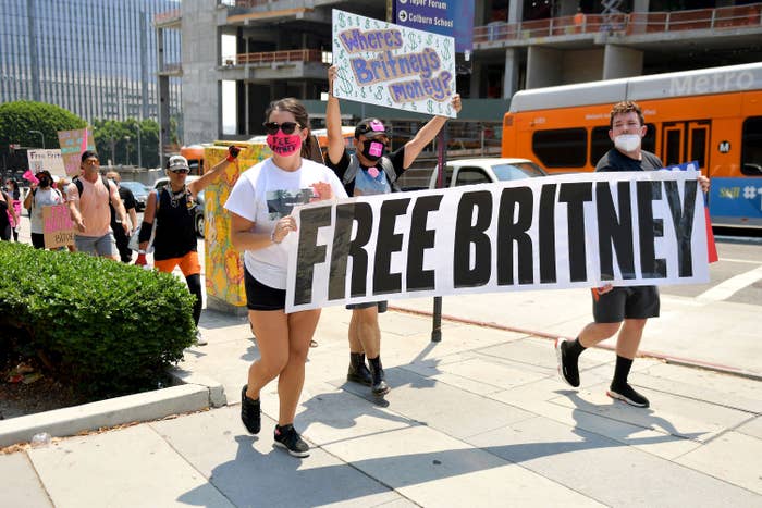 Two protesters in face masks walk with a huge "free Britney" banner stretching across the sidewalk between them; behind them another protester in a face mask holds up a poster reading "Where's Britney's money?" surrounded by dozens of dollar signs