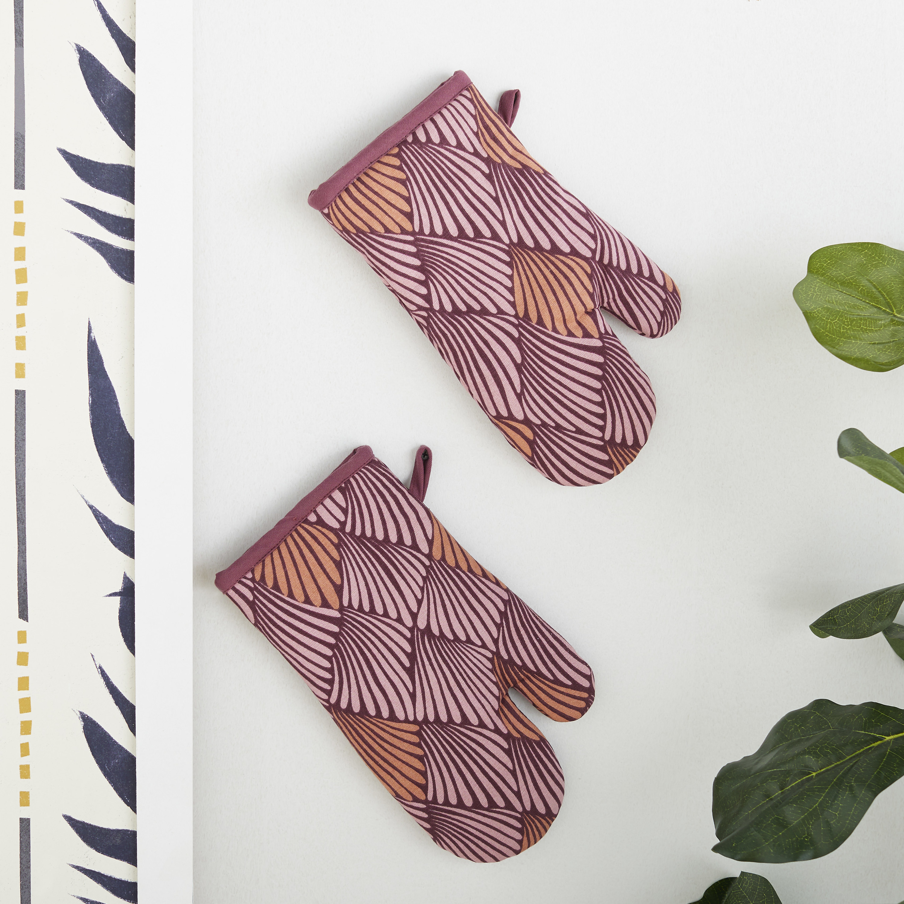 Two oven mitts hanging on the wall with a light pink, orange, and mauve petal-like pattern on them