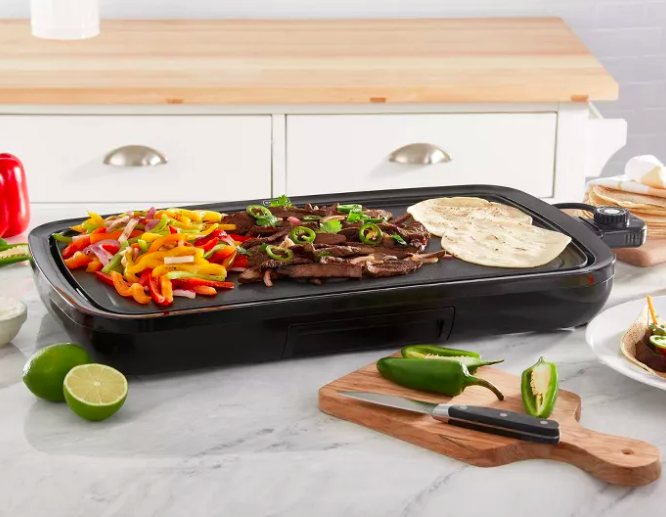 Black griddle with peppers, steak strips, and tortillas on top next to sliced up peppers and limes on a countertop