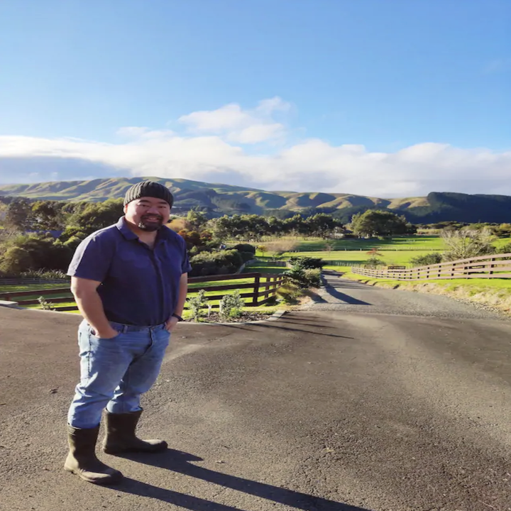 A man in jeans and boots stands smiling on a road leading into a background of lush green grass, mountains, and a bright blue sky