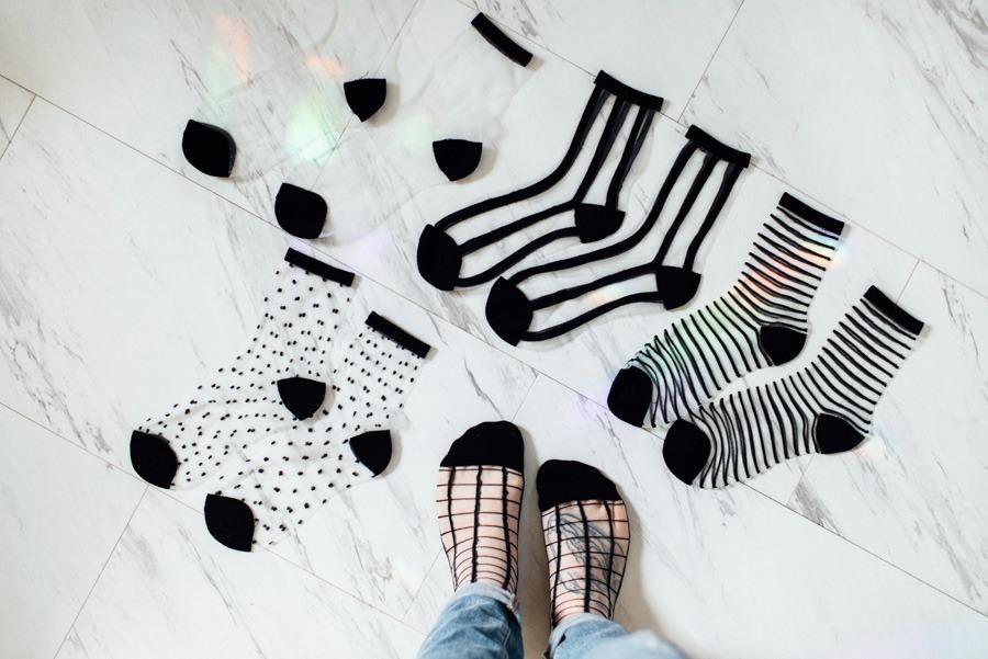 Reviewer pic of four pairs of clear mesh socks laid out on the ground with black toes, heels, and edge with assorted patterns in black, including polka dots, vertical, and horizontal stripes and one pair with checks on the reviewer&#x27;s feet