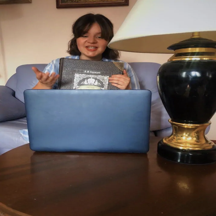 A woman gestures and talks to a laptop while holding a book