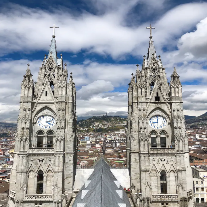 The Basilica of the National Vote towers over the city and into the cloudy sky, its facade gray stone and embedded with a clock face