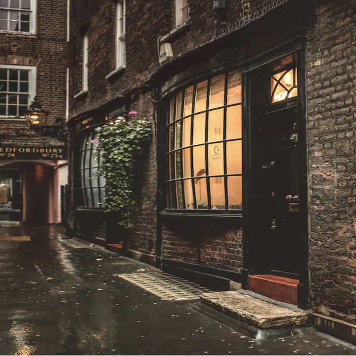 A gloomy, dark alley with brick walls, an old wooden door, and a large window from which warm light appears 