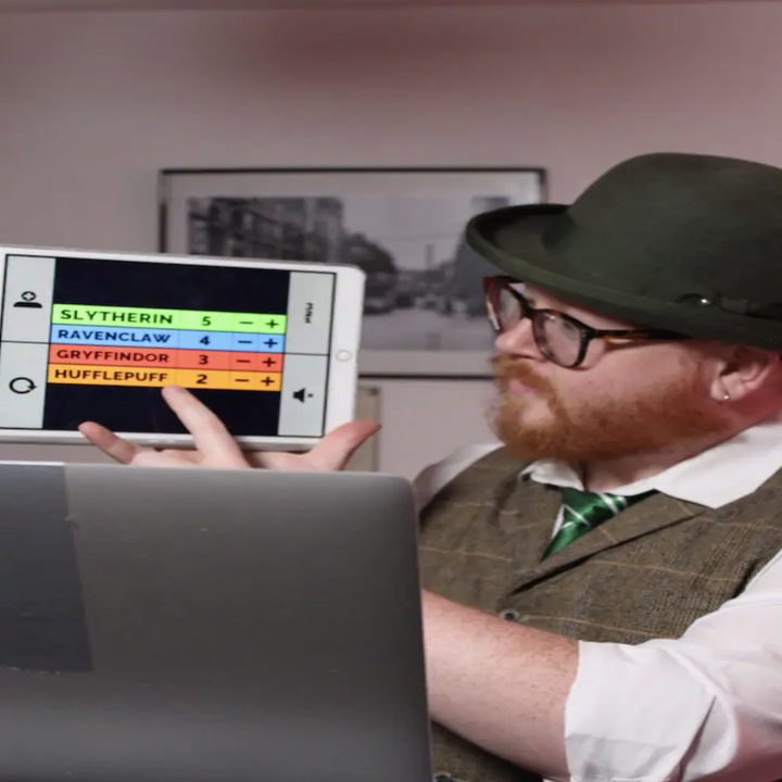A man in a vest, hat, and glasses holds up an ipad to a laptop and points to the screen, which has a chart labeled with the four Hogwarts house names