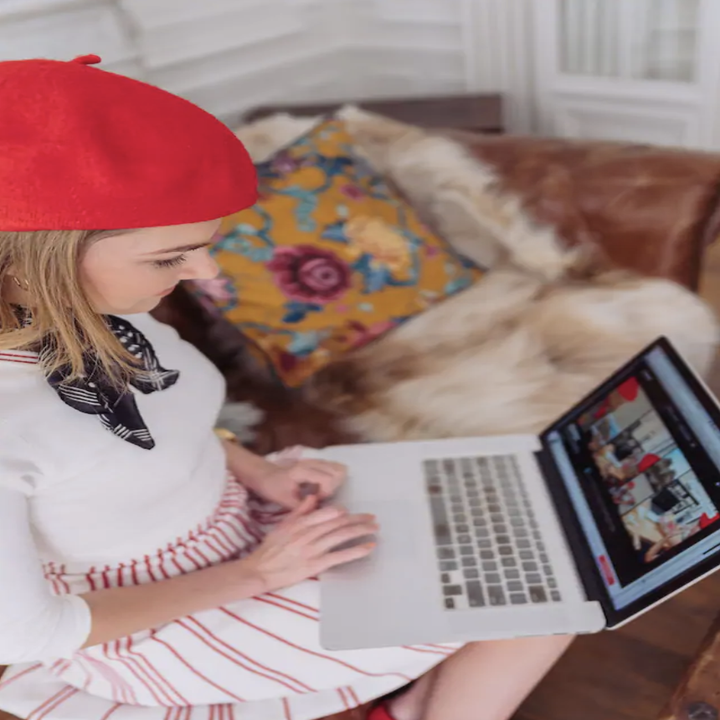 A woman in a red beret sits on a couch and stares down at a laptop, where she is looking at photos