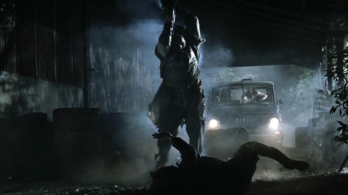 A man in a mask holds a chainsaw over his head while another man is lying on the floor underneath him