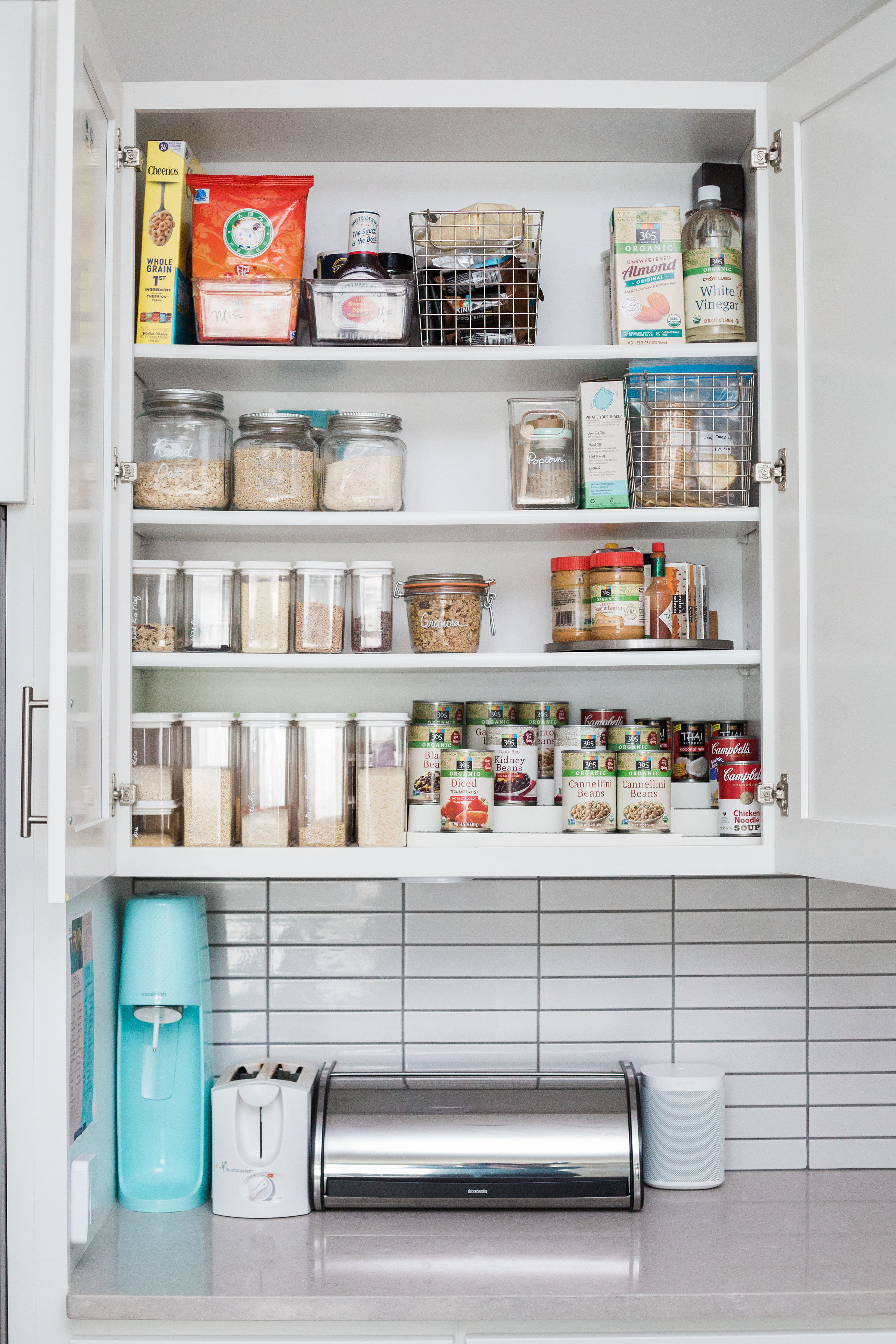 An open cabinet displaying plastic containers and jars filled with staples, a collection of cans on a step shelf, and condiments on a turntable above a Soda Stream, a toaster, a bread saver, and a speaker