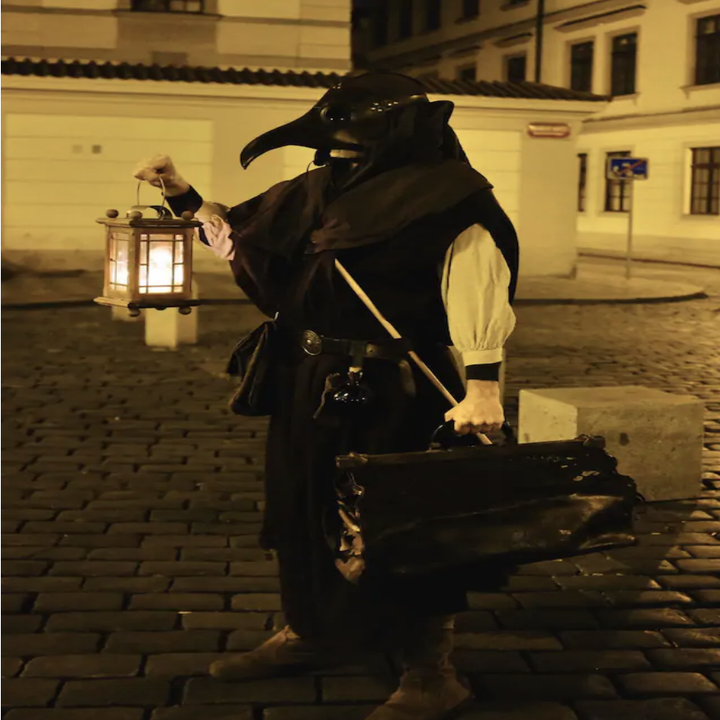 A figure dressed in plague doctor clothes (complete with long, beaked mask) holds a lantern and medicine bag while standing on an empty, cobblestone road