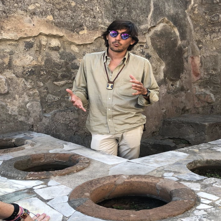 A man gestures and talks to the camera outside in front of a rock wall