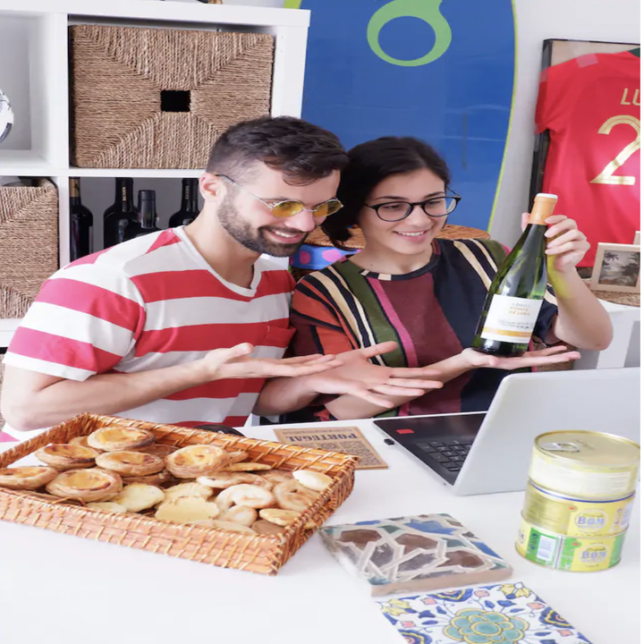 A man and woman gesture and hold up a bottle of wine to a laptop while pastries sit on the table in front of them
